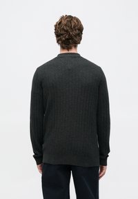 Homem com cabelo castanho ondulado, vestindo um suéter de manga longa cinza escuro texturizado e calças pretas, de costas para a câmara, contra um fundo branco.