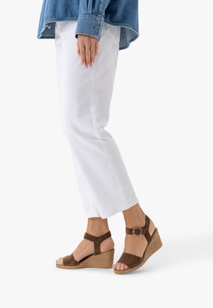 Osoba nosząca białe spodnie i brązowe sandały na koturnie z paskami wokół kostek i złotymi klamrami, stojąca na białym tle.