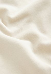 Vaalean kermanvärinen kudottu kangas, jossa on pehmeä pinta ja sileä pinta, esittäen hellävaraisia taitoksia ja hienovaraisia sävyn vaihteluita.