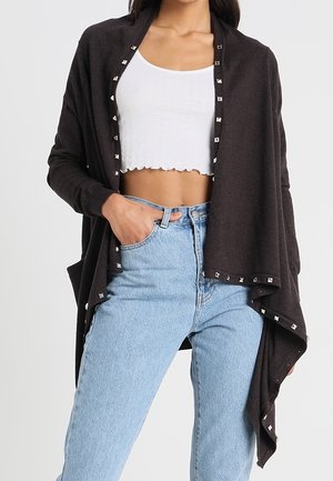 Frau trägt hellblaue High-Waist-Jeans, weißes bauchfreies Tanktop und eine lockere schwarze offene Strickjacke mit silbernen Nieten entlang der Kanten.