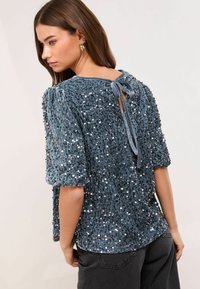 Īsām piedurknēm, teal samta tops, rotāts ar sudraba pērlītēm. Ar sasietu kakla izgriezumu aizmugurē un teksturētu audumu. Izskatās brīvi.