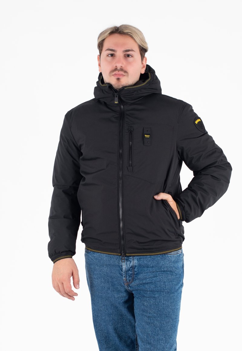 Giacca nera isolata con zipper, cappuccio e tasche frontali. Presenta dettagli gialli e una patch con logo sulla manica. Vestibilità casual.