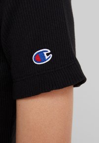 Chemise noire à rayures côtelées à manches courtes avec un petit emblème brodé aux couleurs bleue et rouge sur la manche gauche.