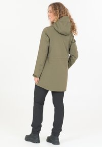 Nainen kiharat hiukset, yllään oliivinvihreä hupullinen takki, mustat housut ja mustat saappaat, seisoo selin ja hymyilee.