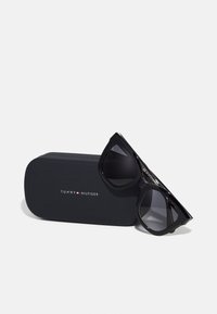 Black sunglasses with dark lenses, angular design, and shiny frame. Includes a black case featuring the Tommy Hilfiger logo.