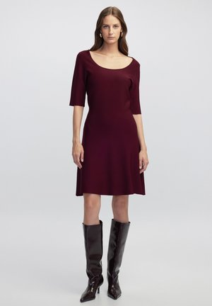 Bordeauxfarbenes Strickkleid mit weitem Rundhalsausschnitt und kurzen Ärmeln, weit auslaufendem Saum, getragen mit knielangen schwarzen Lederstiefeln. Glatte Textur.