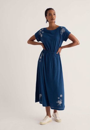 Mujer de pie con las manos en las caderas, vestida con un vestido midi azul con bordado floral blanco y zapatillas blancas, mirando al frente frente a un fondo liso.
