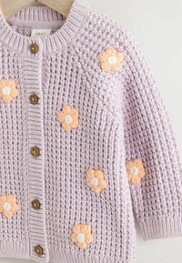 Cardigan bébé tricoté lilas avec boutons marron et appliques de fleurs pêche sur le devant et les manches, suspendu sur un cintre en bois.