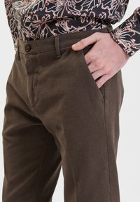 Brązowe spodnie o teksturowanej powierzchni w dopasowanym kroju, z pojedynczym zapięciem na guzik i bocznymi kieszeniami, zestawione z wzorzystą czarną koszulą.