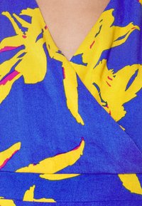 Haut cache-cœur col en V avec un fond bleu orné de motifs floraux jaunes vifs et des accents rouges. Tissu lisse avec une texture douce.