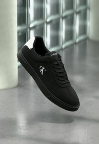 Baskets noires Calvin Klein avec languette talon blanche, lacets noirs et logo CK sur le côté, présentées devant un mur carrelé brillant.
