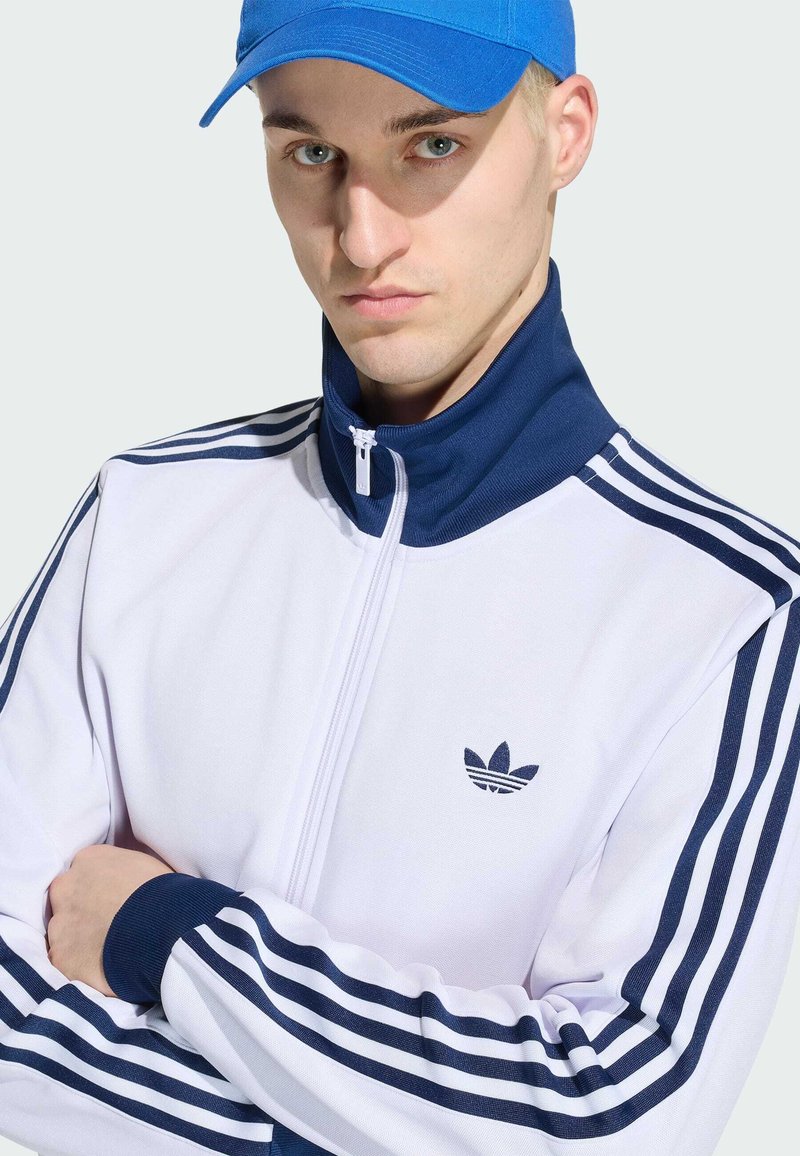 Hombre con gorra azul y chaqueta de chándal blanca de Adidas con rayas azul marino, brazos cruzados, mirando hacia adelante sobre un fondo liso.