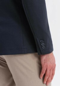 Närbild av en persons hand som bär en mörk marinblå kavajsleeve med tre knappar, ihop med beiga byxor.