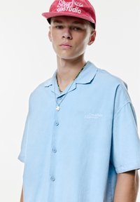 Cămașă bleu deschis, cu mânecă scurtă, tip cămașă cu nasturi, cu o croială lejeră și broderie "Creative Manner". Modelul poartă o șapcă roz și un colier.