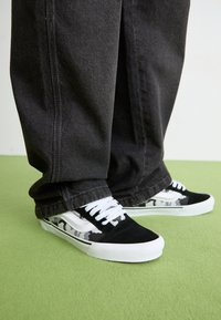 Schwarz-weiße Sneakers mit einem Camouflage-Muster, ausgestattet mit einem Obermaterial aus Wildleder, weißen Schnürsenkeln und einer weißen Gummisohle, kombiniert mit dunklen Hosen.