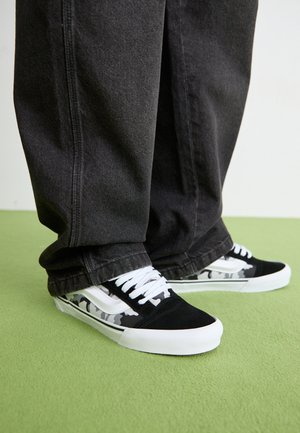 Black and white sneakers with a camouflage pattern, featuring a suede upper, white laces, and a white rubber sole, paired with dark pants.