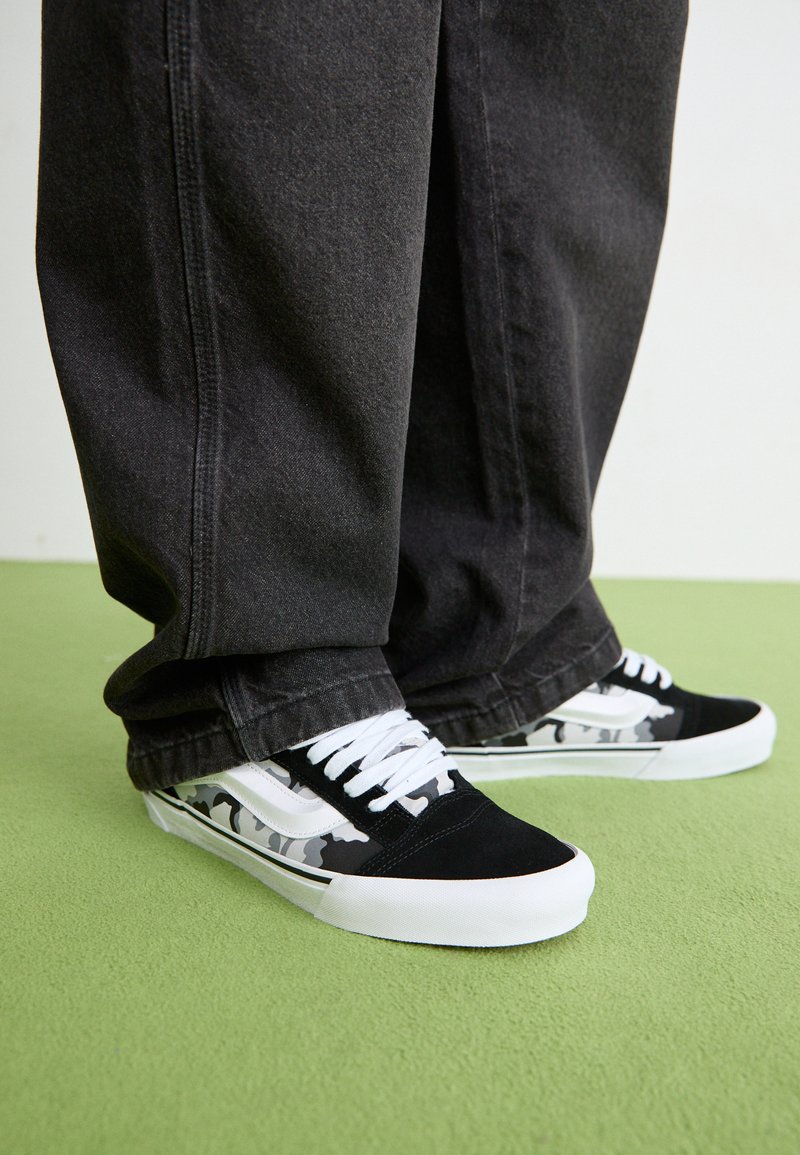 Schwarz-weiße Sneakers mit einem Camouflage-Muster, ausgestattet mit einem Obermaterial aus Wildleder, weißen Schnürsenkeln und einer weißen Gummisohle, kombiniert mit dunklen Hosen.