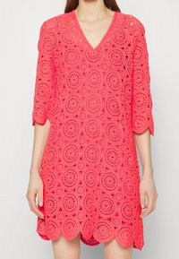 Vestido de ganchillo color coral con escote en V, mangas tres cuartos y patrones circulares. El dobladillo festoneado añade detalle a la tela texturizada.
