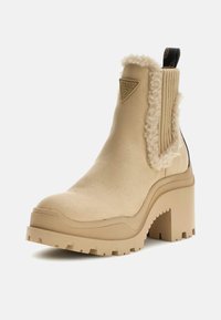 Bota beige hasta el tobillo con un borde texturizado de pelo sintético y paneles laterales elásticos. Tacón grueso y robusto y suela rugosa para mayor tracción. Detalle del logo en forma de triángulo.