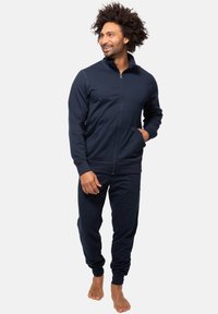 Marinefarbener Zip-Hoodie und dazu passende Hose aus weichem Stoff. Mit Seitentaschen, elastischen Bündchen und einem Kragen. Das Model ist barfuß.