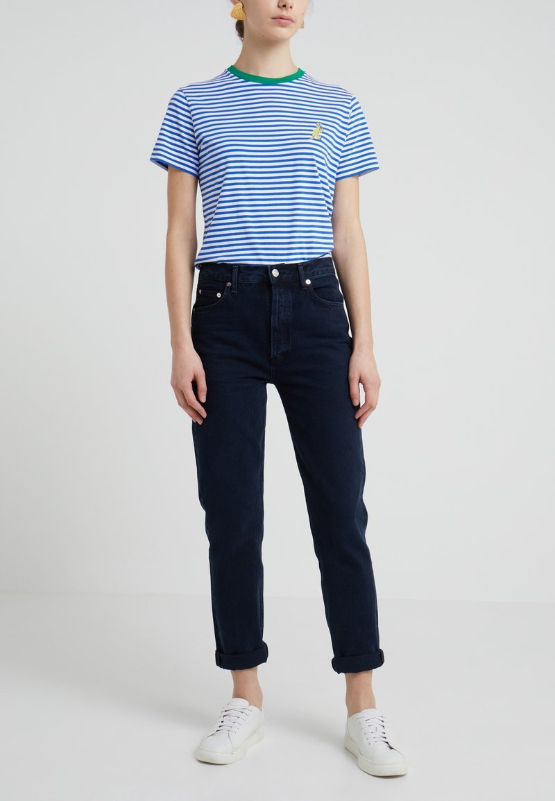 Personne portant un t-shirt rayé bleu et blanc avec col vert, un jean foncé retroussé, des baskets blanches et des boucles d’oreilles jaunes, se tenant devant un fond uni.