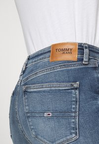 Vaqueros azules con un acabado texturizado, que cuentan con una etiqueta de cuero etiquetada "TOMMY JEANS" en la cintura y un pequeño logo en el bolsillo trasero.