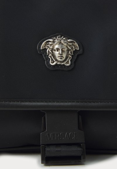 Silver Medusa emblem on black fabric above a black clasp with "VERSACE" embossed on it, part of a bag or accessory.