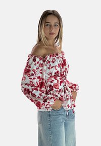 Blusa bianca scollata con stampa floreale rossa, colletto arricciato elasticizzato e vita stretta. Abbinata a jeans azzurri larghi.