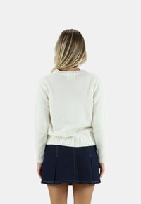 Biały sweter z dzianiny z prążkowanym dołem i mankietami, w parze z ciemnoniebieską spódnicą w plisy. Modelka widoczna z tyłu.