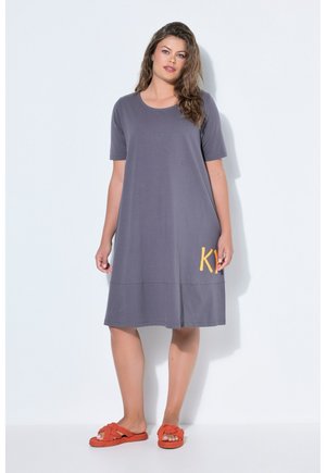 Femme portant une robe grise ample allant jusqu'aux genoux avec des manches courtes et des chaussons orange, debout devant un fond blanc uni.