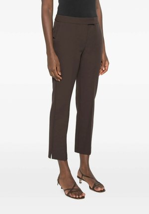 Vrouw draagt donkerbruine cropped broek met kleine zijsplitten gecombineerd met zwarte sandaaltjes met lage hak en bandjes tegen een witte achtergrond.