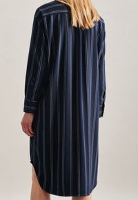 Mörkblå skjortklänning med vertikala vita ränder, långa ärmar, knäppte manschetter och sidoklyftor. Tillverkad av ett slätt, mjukt tyg.