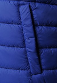 Modrá péřová bunda zblízka, s prošívanou texturou, horizontálním šitím a boční kapsou integrovanou do designu. Hladký povrch látky.