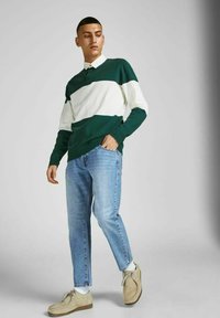 Jersey de polo de punto a rayas verdes y blancas, jeans azules, zapatos beige, con un ajuste relajado y una textura sutil. El modelo se presenta de manera casual.