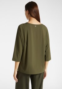 Blusa leggera verde oliva con maniche a tre quarti e scollatura rotonda. Presenta un piccolo bottone decorativo sul retro.