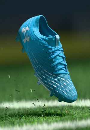 Blauwe voetbalschoen met een gestructureerd bovenwerk, voorzien van een geometrisch patroon en lichtblauwe veters, hangend boven gras met vuildeeltjes.