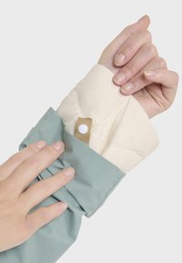 Le poignet d'une veste bleu clair révèle une doublure crème, dotée d'une fermeture à bouton-pression beige. Le tissu semble lisse et résistant à l'eau.