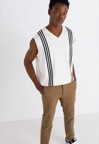 White sleeveless top with black vertical stripes. V-neck design. Paired with brown pants and black sneakers. Plain background.