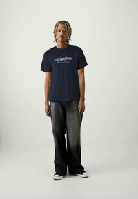 Ungdom med flettehår iført en marineblå t-skjorte med grafikk, falmede vide jeans og svarte lave sneakers står mot en ensfarget bakgrunn.