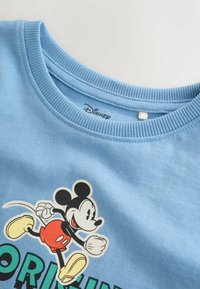 Chemise bleu clair avec graphique de Mickey Mouse en train de courir et texte partiel, col rond côtelé, étiquette Disney à l'intérieur de l'encolure.