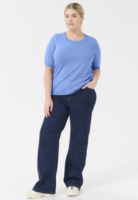 Camisa de manga corta azul claro con cuello redondo, combinada con jeans rectos de pierna oscura. El conjunto se complementa con zapatillas de color claro.