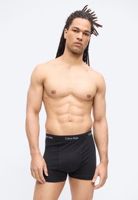 Muskuløs mand med lange dreadlocks iført sorte Calvin Klein boxer-briefs, stående mod en ensartet lys baggrund.