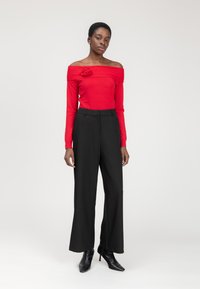 Rode off-shoulder trui met bloemdetail, gecombineerd met hoge zwarte wijde broek en zwarte puntige hakken. Effen achtergrond.