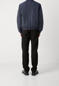 Granatowa kurtka bomberka z elastycznymi mankietami i ściągaczem u dołu, zestawiona z czarnymi spodniami i ciemnym obuwiem, widziana od tyłu. Gładka faktura tkaniny.