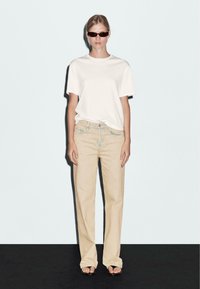T-shirt bianco a maniche corte abbinato a jeans beige a gamba larga. Il modello indossa occhiali da sole scuri e sandali aperti. Design semplice, colori neutri.