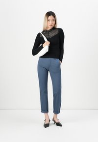 Svart långärmad topp med spetsdesign, blå uppruffade byxor och svarta spetsiga klackar. Modellen håller en liten vit väska.