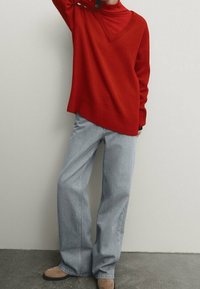 Pull en tricot rouge avec un col diagonal, associé à un jean large bleu clair et des bottines beiges. Texture lisse, coupe décontractée.