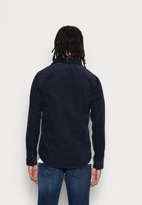 Camisa de manga larga marina en tejido suave, con cuello abotonado, canesú trasero y bajo redondeado, combinada con jeans azules.