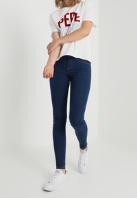 Camiseta blanca con texto rojo y azul, combinada con jeans skinny azul oscuro y zapatillas blancas, con un diseño ajustado y una textura suave.
