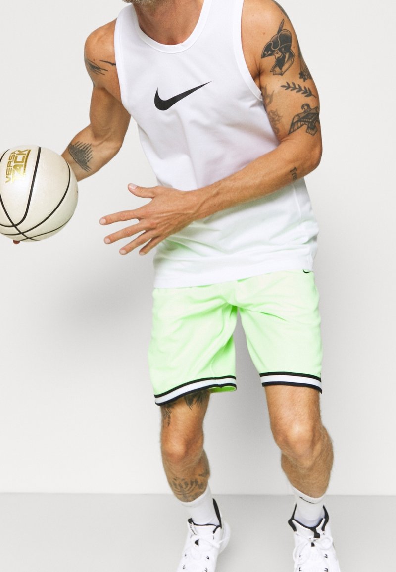 Camiseta sin mangas blanca de Nike con logo negro, combinada con pantalones cortos verdes neón con ribete negro y un balón de baloncesto en la mano.